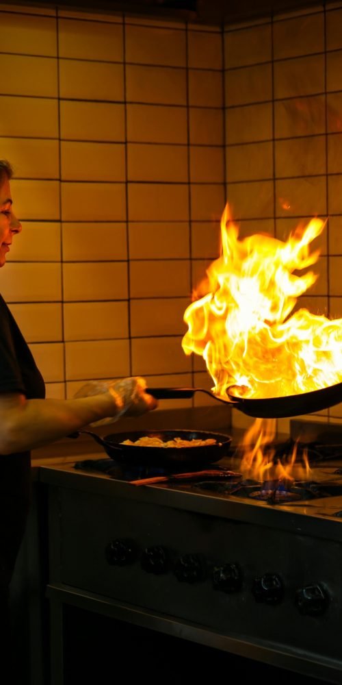 A female chef expertly handles a frying pan as flames rise, showcasing cooking skills in a professional kitchen.
